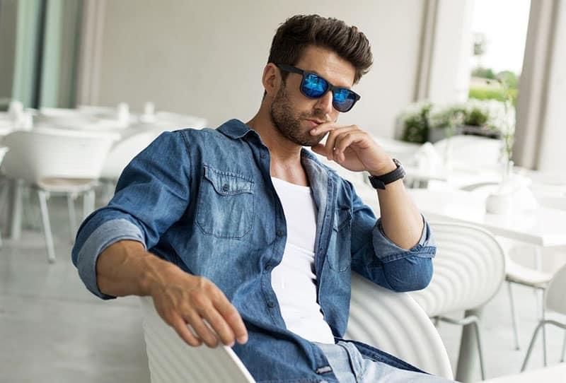 guy sitting on white chair wearing a denim jacket and sunnies