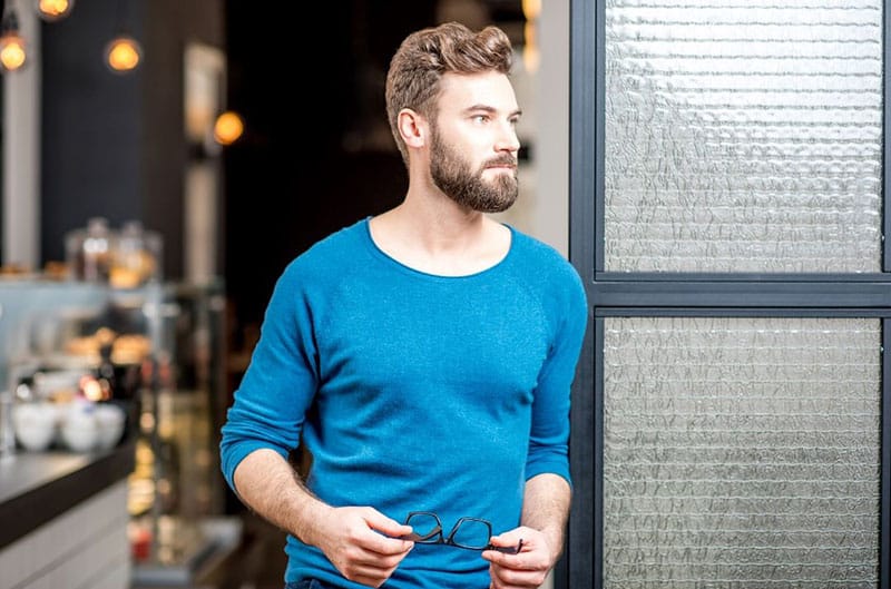 um homem bonito com um top azul, parado à porta, segurando um óculos