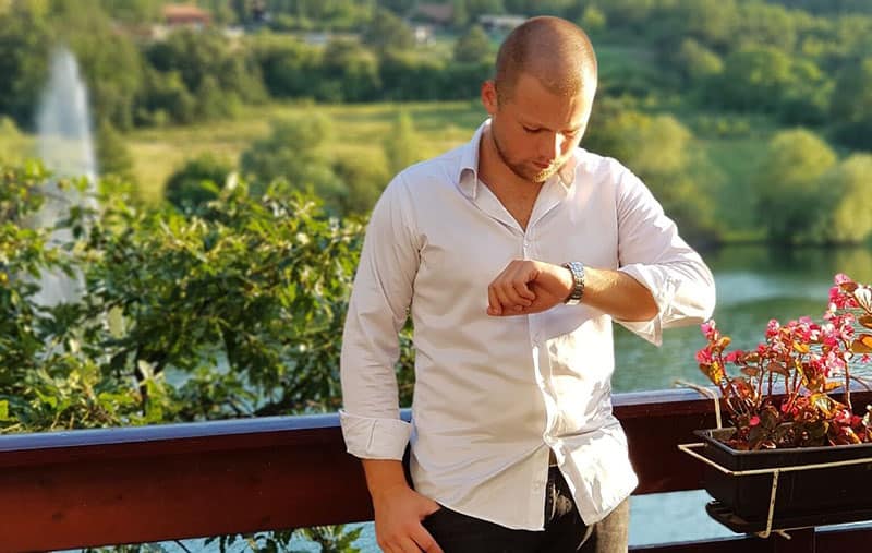 hombre guapo de pie mirando su reloj apoyado en la barandilla cerca de una masa de agua