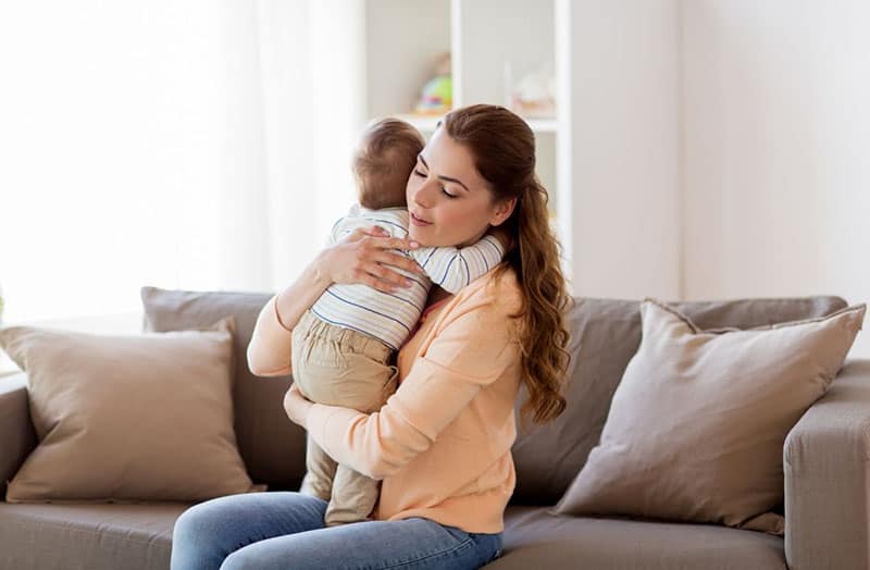 happy young mother holding her toddler in her arms hugging her while sitting on a sofa inside the living room
