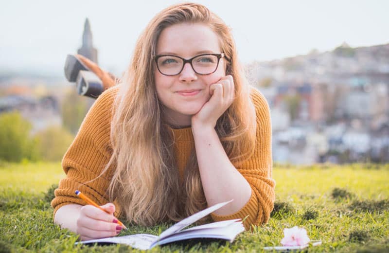 mujer joven feliz que escribe en su cuaderno mientras que se pone en cuclillas en la tierra