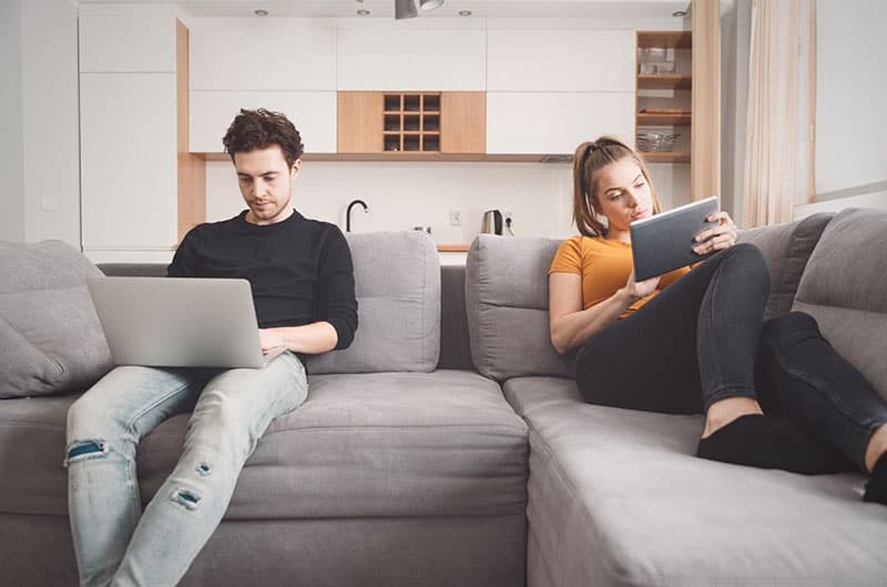 indifferent couple sitting on sofa far from each other facing their own gadgets 