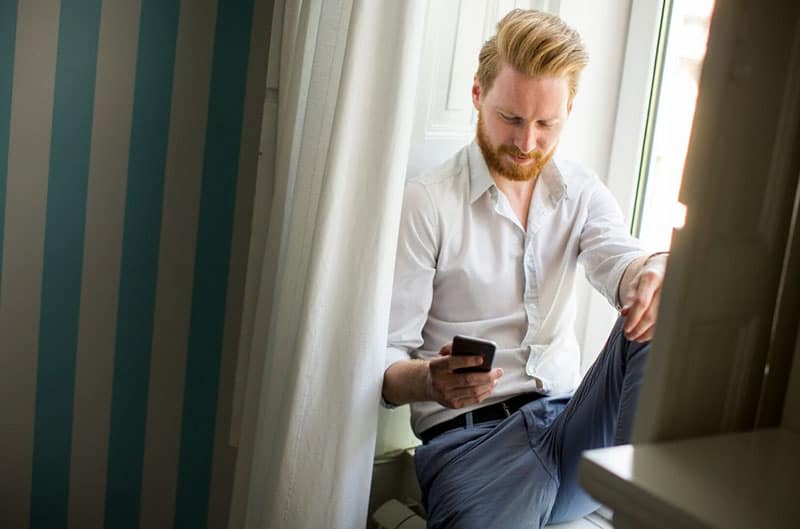 man holding a smartphone sitting on the window sill partly covered with curtains