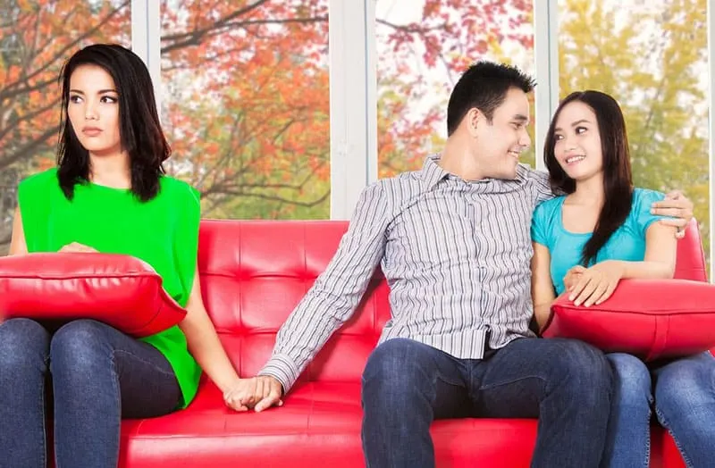 man holding hand of a woman while his other arms is wrapped around another woman sitting on the red sofa