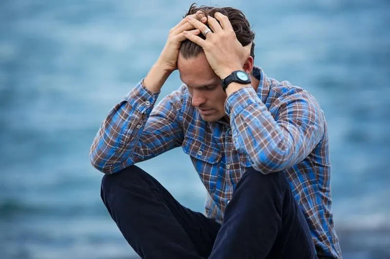 man in checkered sitting near a body of water with hands on his head feeling sad 