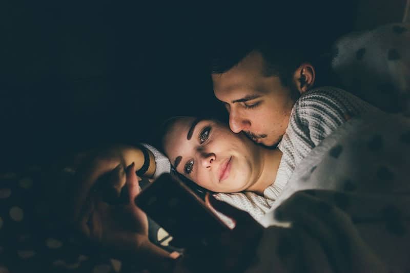 man kissing the woman in bed while the woman is busy on her smartphone