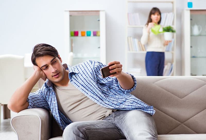 hombre apoyado en el sofa mirando su smartphone con mujer mirandolo regando plantas