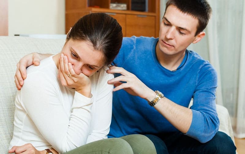 man sitting next to a crying woman sitting in a sofa inside living room