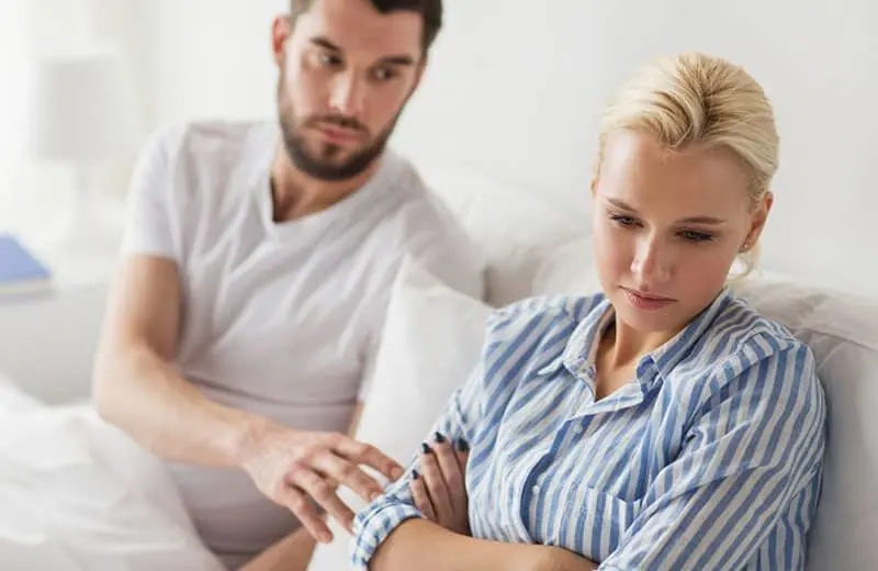 man touching the sad woman sitting in the sofa wearing while and blue top