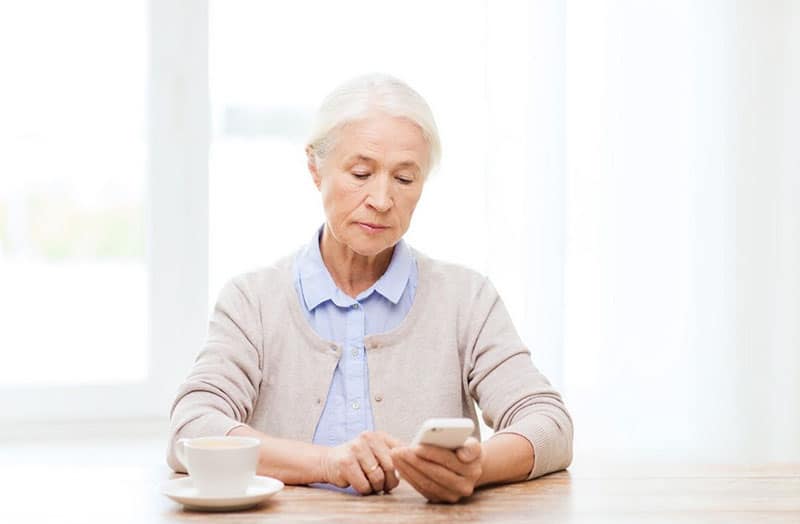 old woman dialing on a smartphone while sitting with cup on the table
