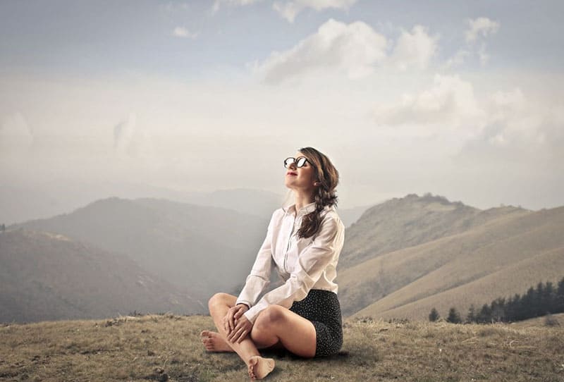 mujer feliz pacífica que se sienta en la tierra en la tapa de la montaña descalza que usa el traje ocasional con los anteojos