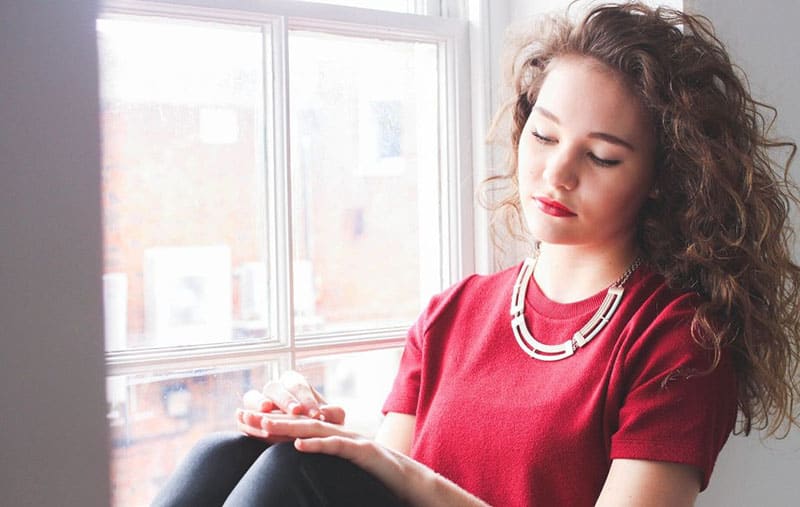 pensive curly haired woman sitting in the window sill wearing red top