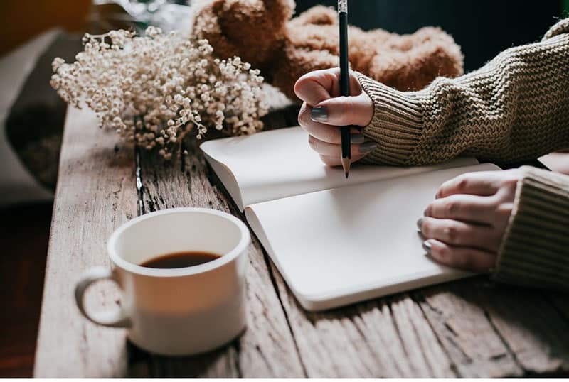 person's hand writing on a notebook placed on the table with a cup of coffee at the side