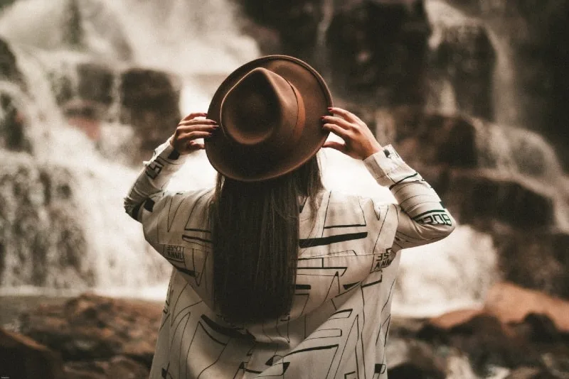 woman holding brown hat while looking at waterfall