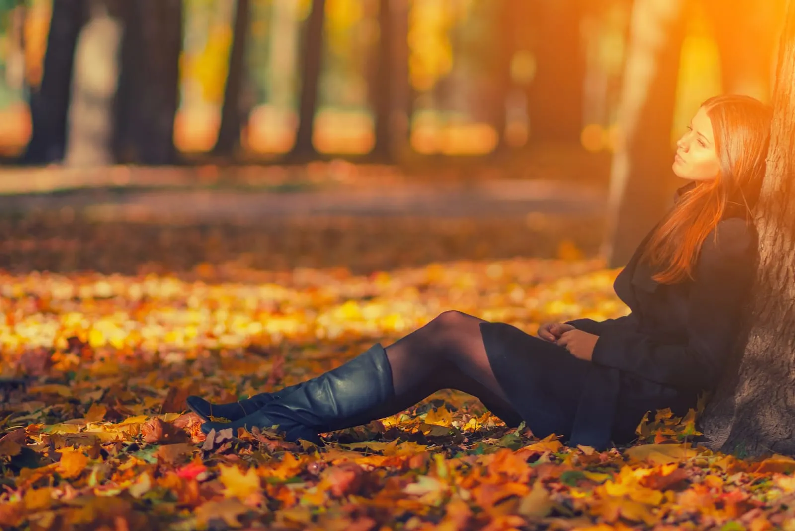 woman in black coat and leather boots sitting under tree in autumn park
