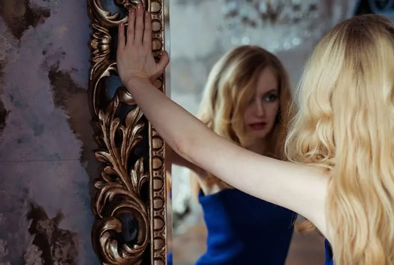 woman in blue dress facing the mirror with hands on the edges of the mirror
