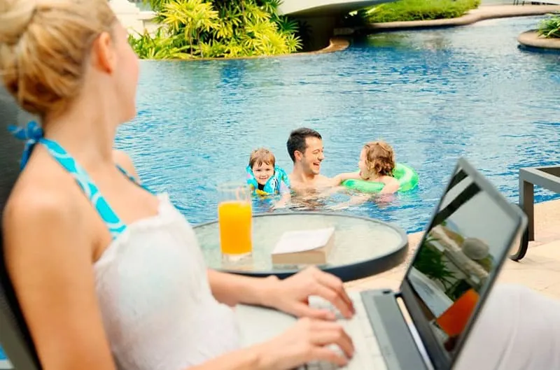 woman looking at her family enjoying at the pool while she is on her laptop