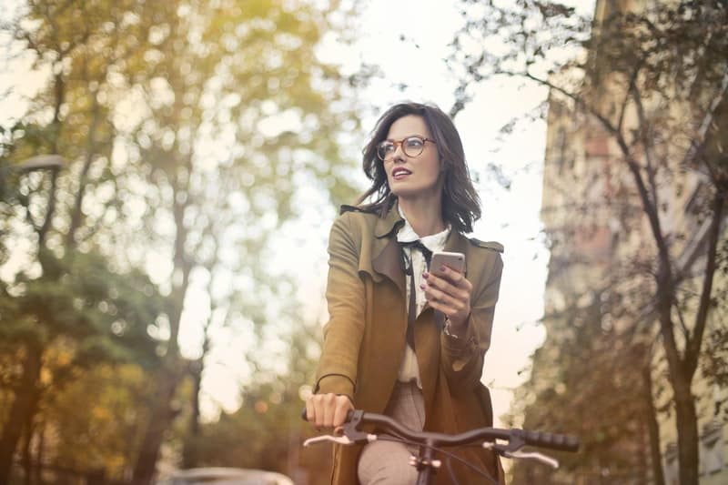 mujer montando en bicicleta mientras sostiene un smartphone en medio de la carretera con muchos árboles