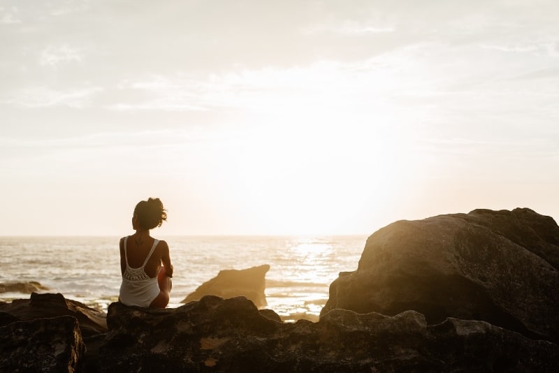 woman in white swimsuit sitting on stone near ocean