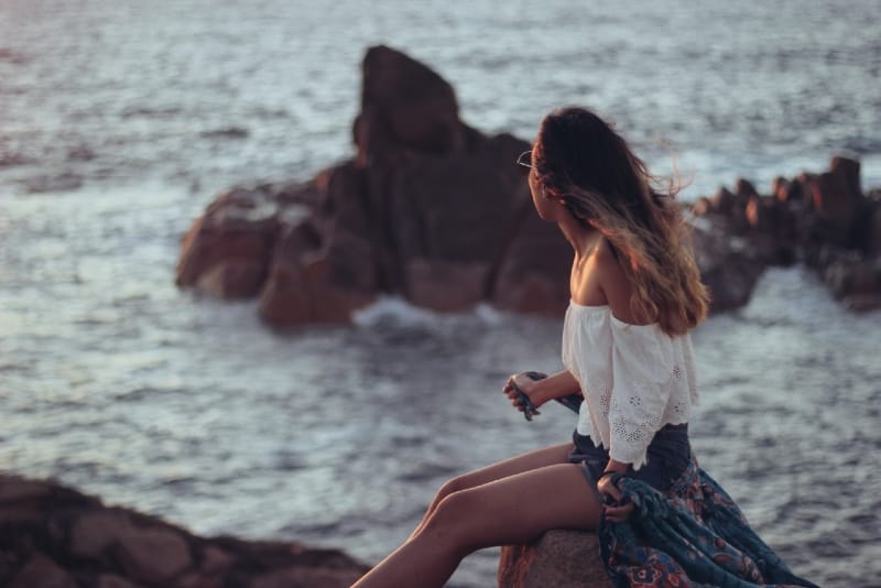 woman in white top sitting on stone looking at sea