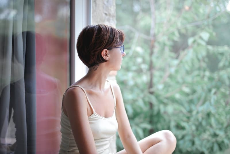mujer con top blanco sentada en el cristal de una ventana mirando al exterior
