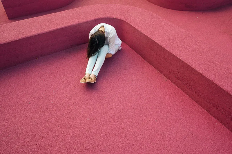 woman tucked in a corner of a pink structure wearing white top and jeans with head bowed down