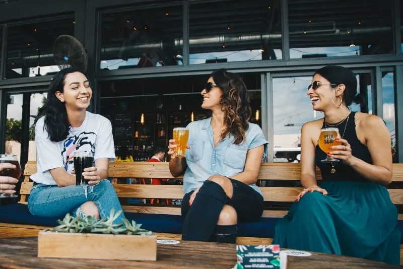 three smiling women sitting on bench holding glasses of beer