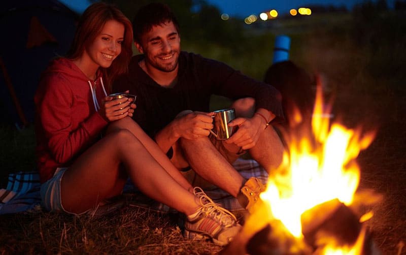 young couple during summer night sitting drinking tea in front of a bonfire