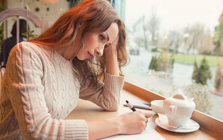 woman writing on a paper inside a cafe with a tea pitcher on the table