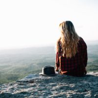 blonde woman sitting on cliff with hat by its side