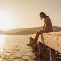 mulher jovem e bonita sentada num cais de um lago a olhar para a água ao pôr do sol