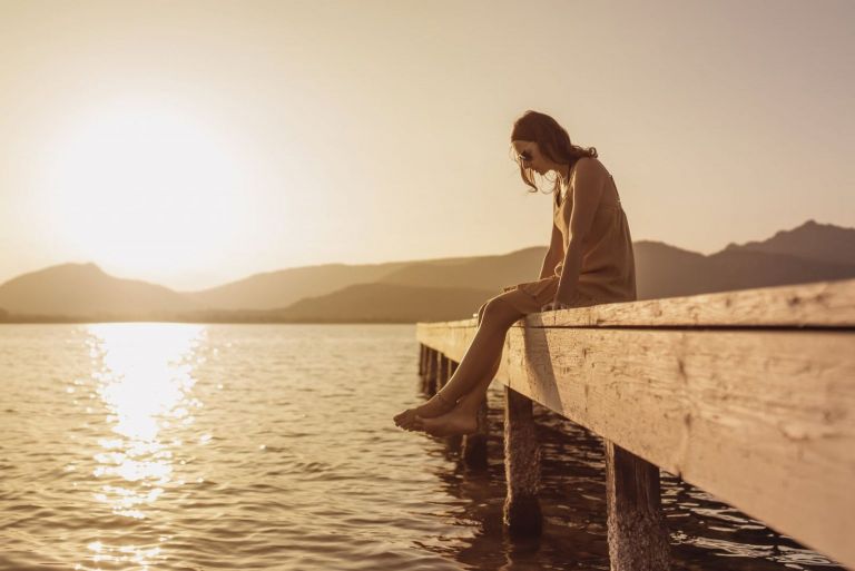 pretty young woman sitting on a pier of a lake looking down to the water at sunset