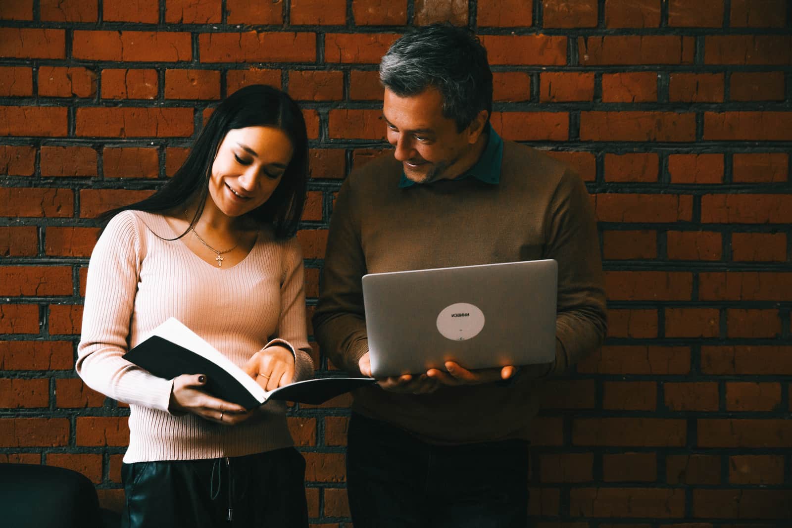 man and woman looking at notes researched on the laptop while standing near brick walls