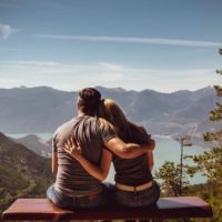 man and woman sitting next to each other looking at the lake and mountains in front of them