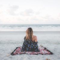 mulher sentada numa manta na praia a olhar para o oceano