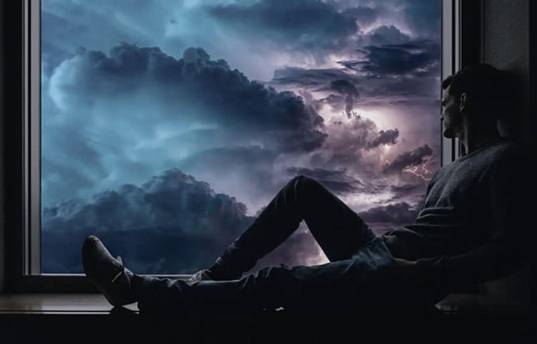 man sitting on the window sill looking outside thru a stormy weather