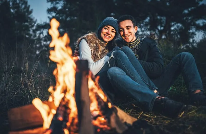 couple dating with bonfire in front of them sitting on the ground in the woods on golden hour
