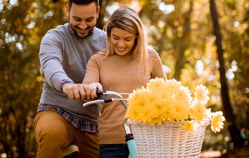 couple sharing a bike with a basket of flower in the park