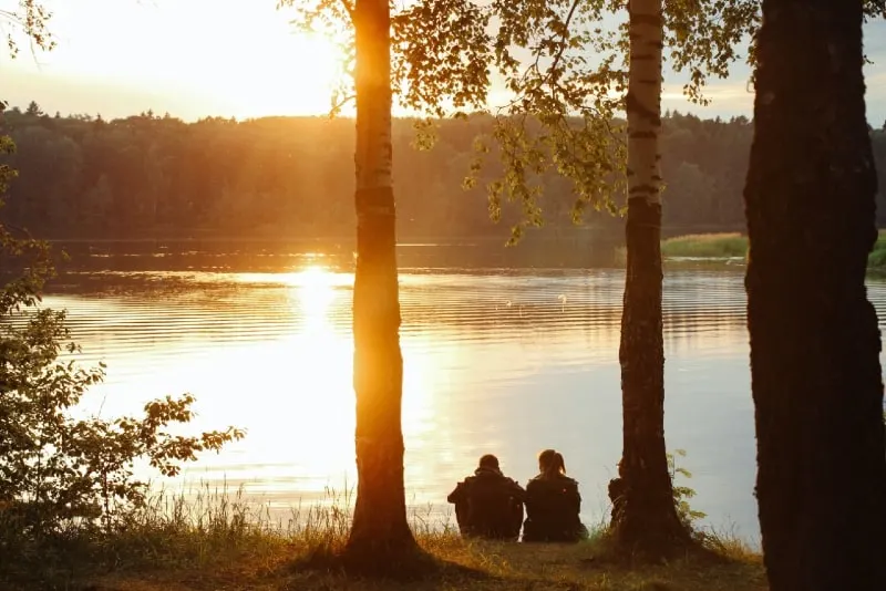 man and woman sitting on grass near water during sunset