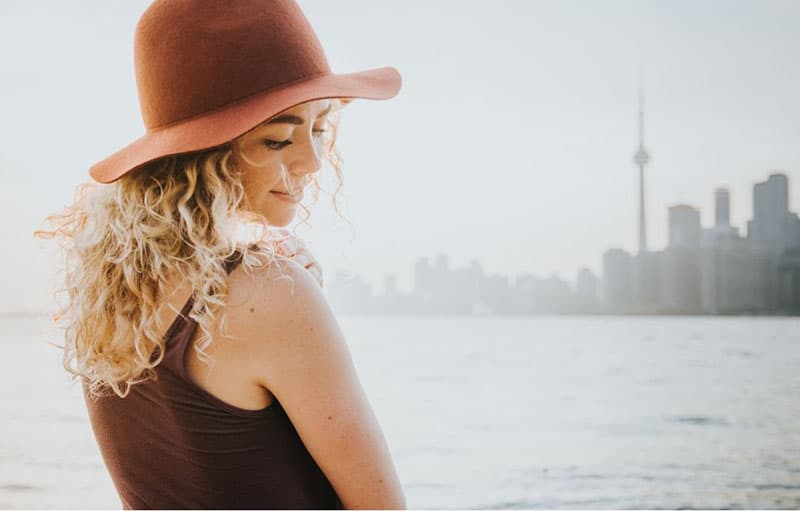 curly haired woman with a hat smiling and standing near a body of water near the city
