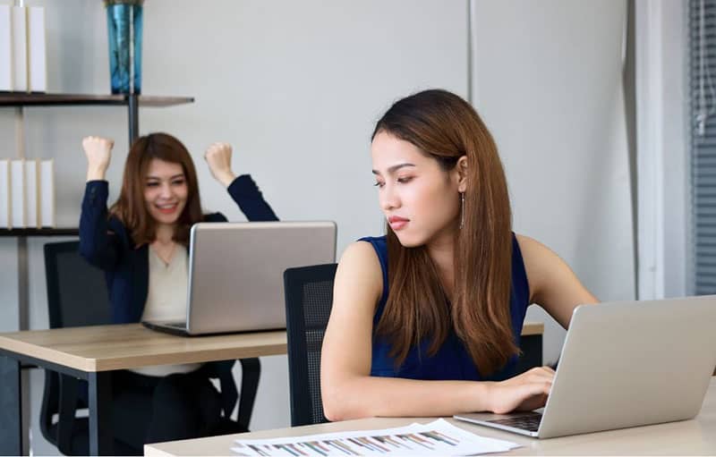 envious angry woman looking at her victorious colleague seated at her back