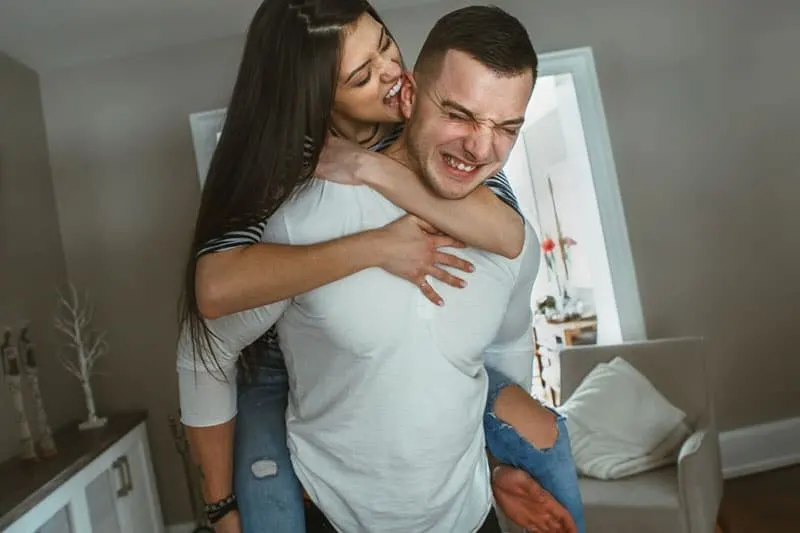 happy funny couple inside living room, woman biting and piggy back on the man