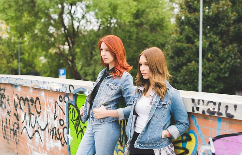 figura in ginocchio di due donne che indossano giacche di jeans e jeans appoggiate al muro