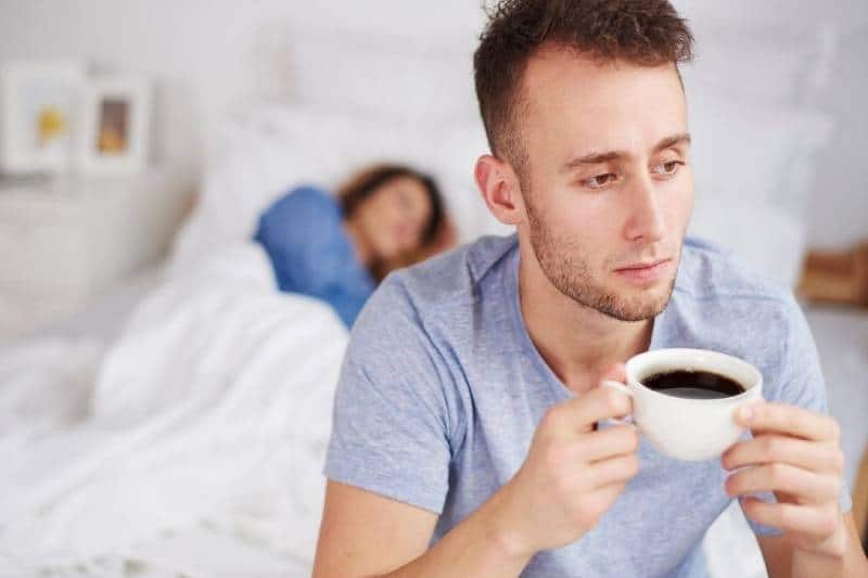 man drinking coffee in bed and a woman sleeping behind him