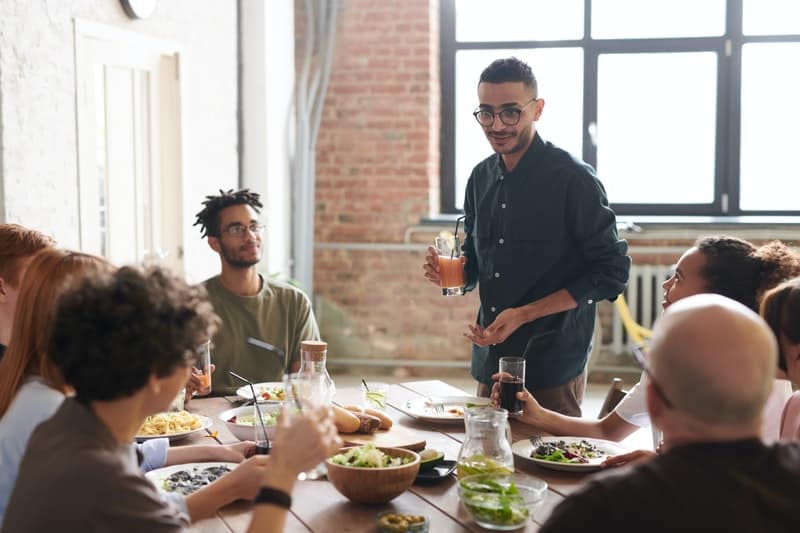 man holding a drink standing in the middle of people dining and delivering a speech