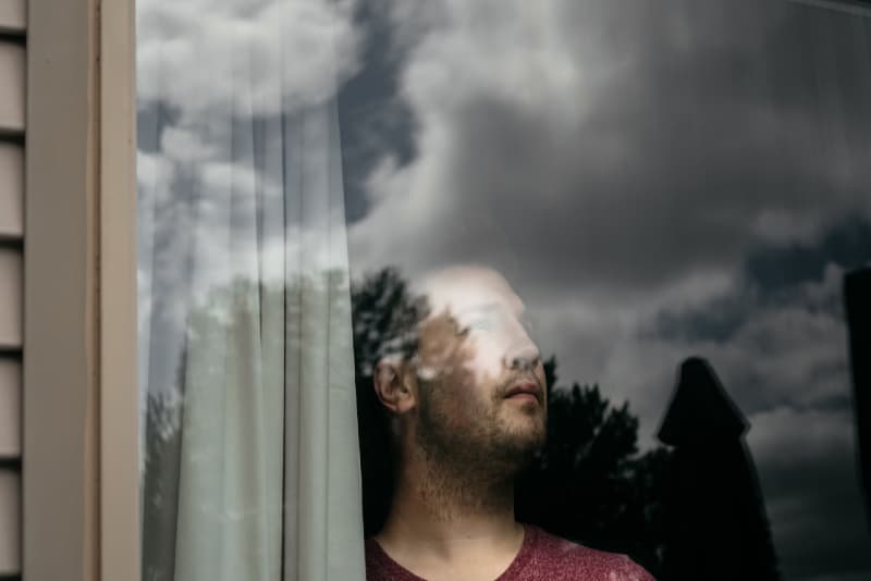 man looking through window while standing near white curtain