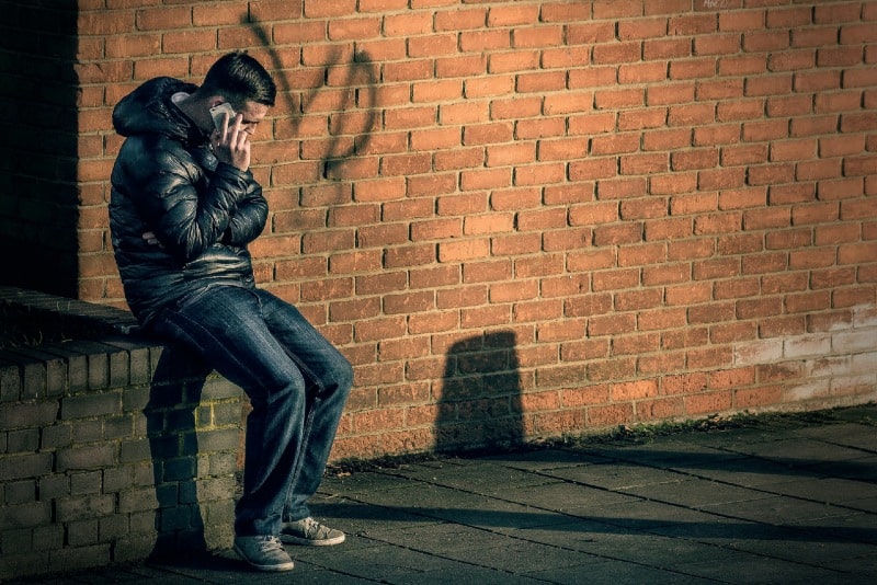 homem a falar ao telefone enquanto está sentado numa parede de tijolo