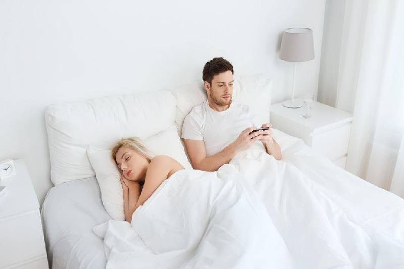 man texting in bed seated next to a sleeping woman