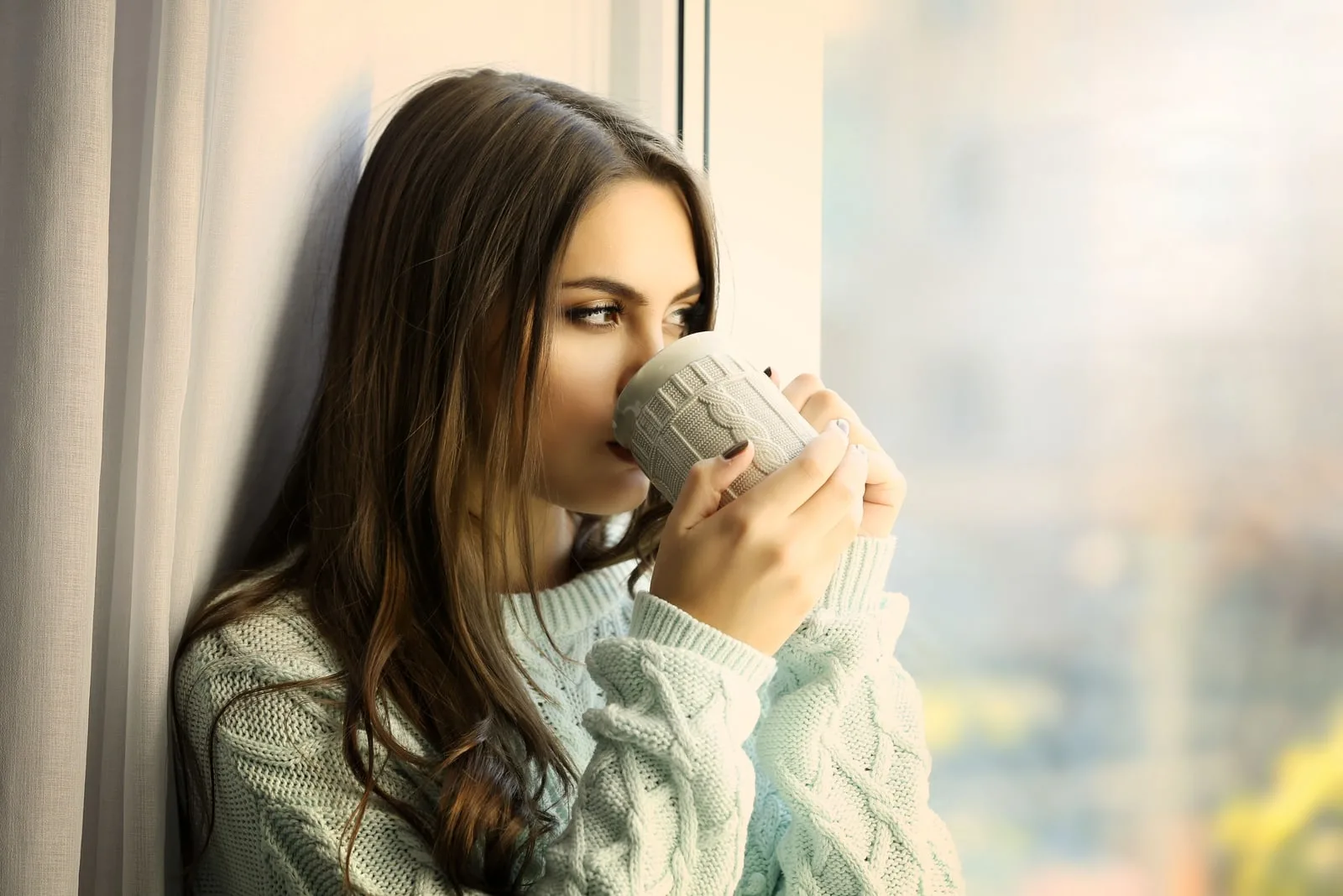 pensive brunette woman drinking coffee by the window