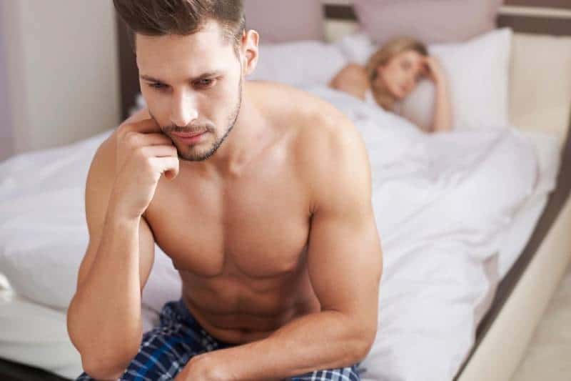 pensive half naked man sitting on bed and a woman sleeping on bed